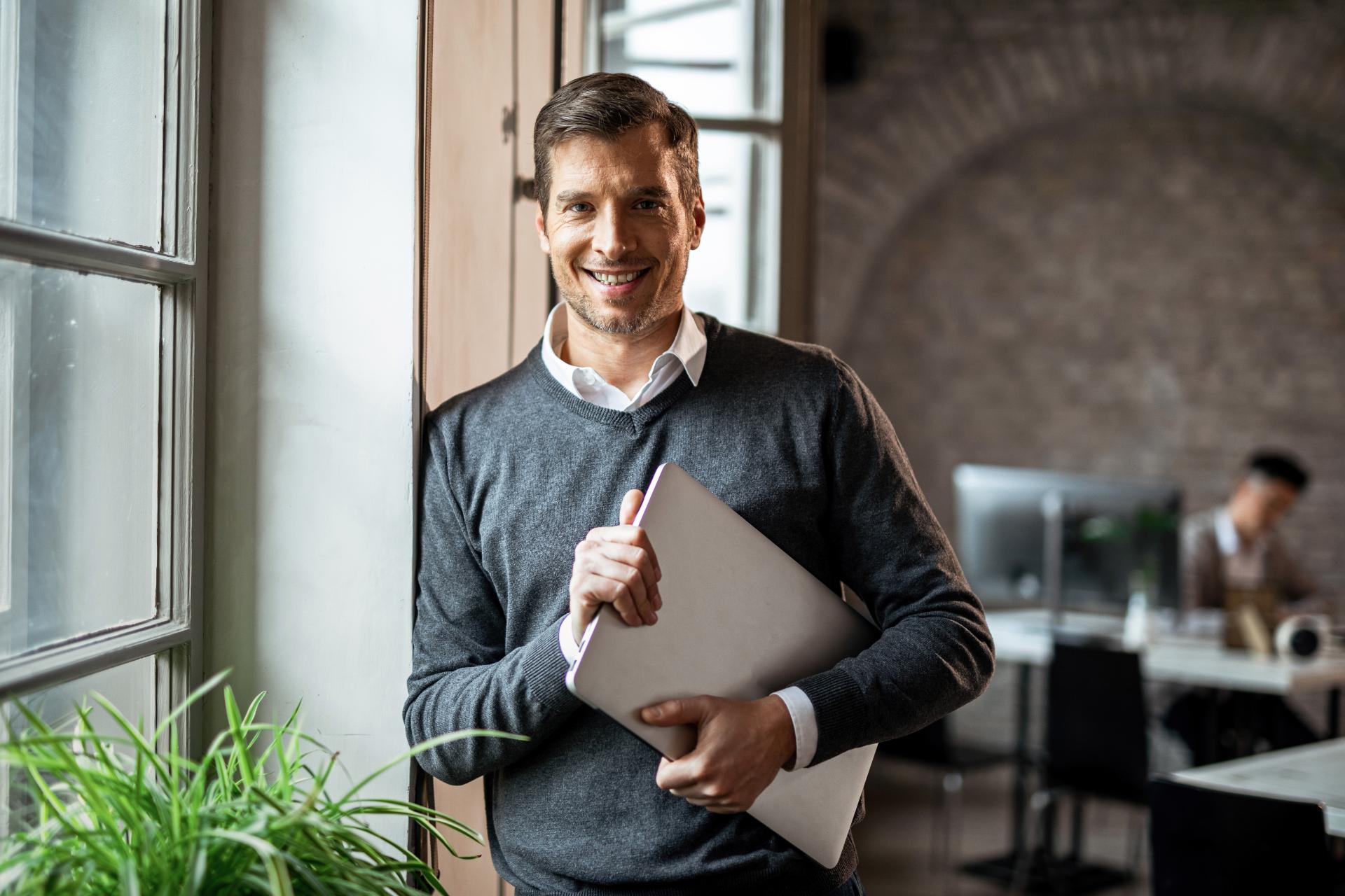 Happy Freelance Worker With Laptop Looking Camera While Standing By Window Office